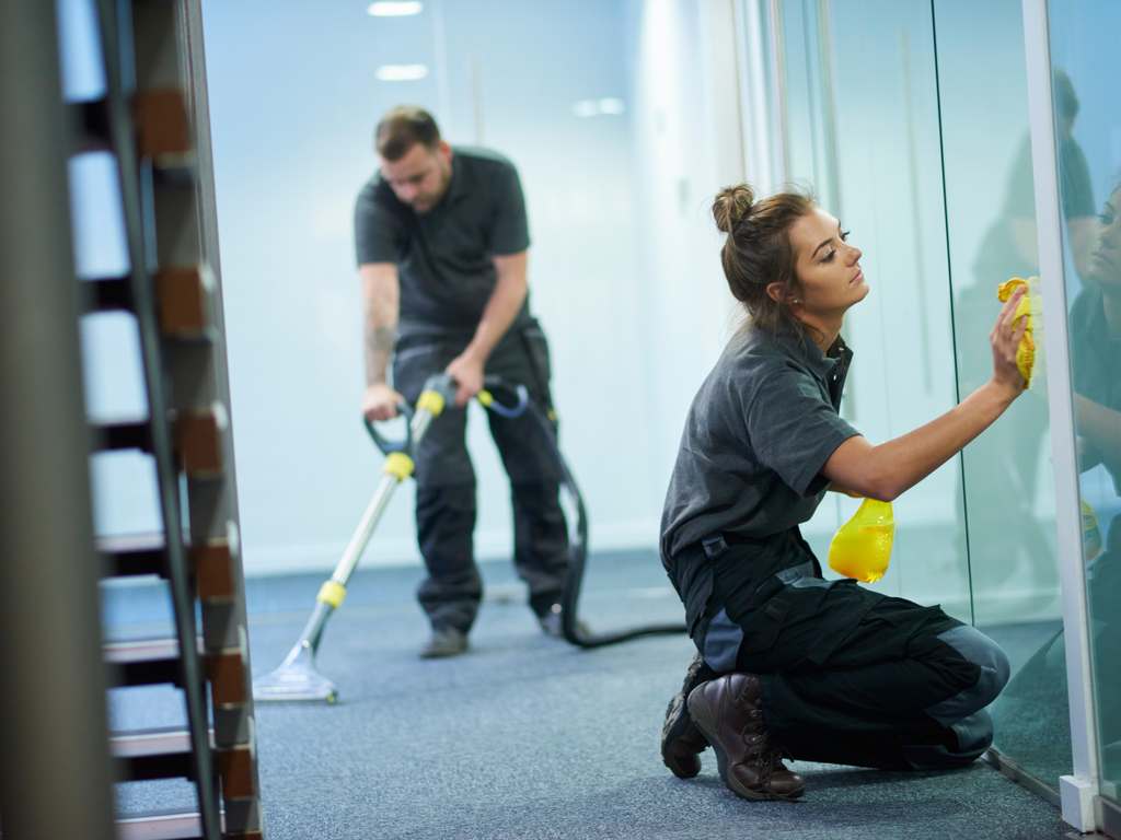A professional commercial cleaning company employee wipes a glass partition while another operates a floor machine in the background of an office.