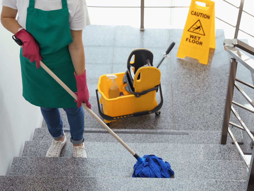 A person in a green apron and pink gloves mops a staircase near a yellow bucket and wet floor sign as part of professional commercial cleaning services in Marietta, Georgia.