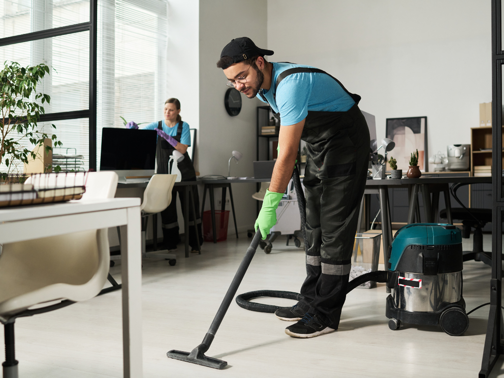A cleaner uses a shop vacuum on an office floor while another worker cleans a desk, helping to lower the office cleaning cost in Marietta.